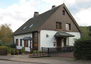 Hausansicht.jpg Ein zweistöckiges, weißes Haus mit dunkelbraunem Walmdach und weißen Holzfenstern in ruhiger Lage.