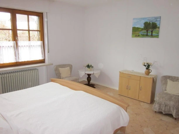 Schlafzimmer Helles Schlafzimmer mit Doppelbett, zwei Sesseln, hellem Holzschrank und Fenster mit weißem Vorhang.