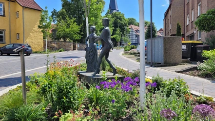 Bronzeskulptur eines tanzenden Paars inmitten eines bunt blühenden Blumenbeets vor Fachwerkhäusern.
