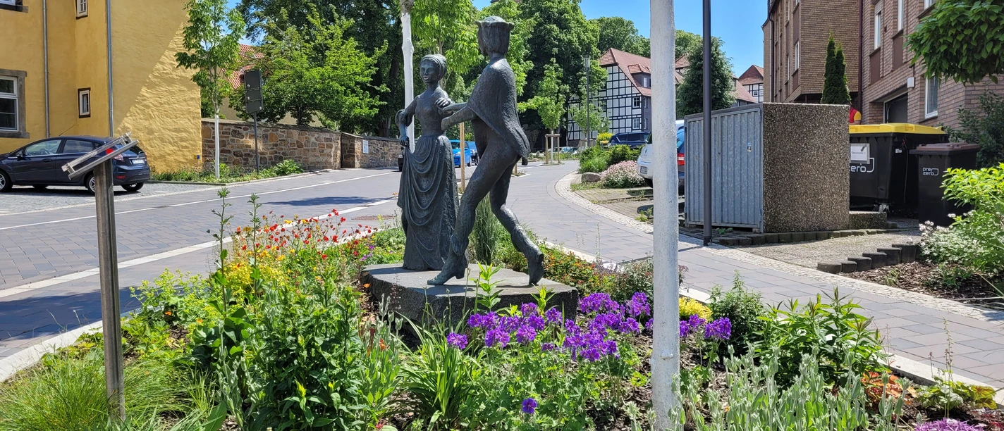 Danzelstätte Lübbecke Bronzeskulptur eines tanzenden Paars inmitten eines bunt blühenden Blumenbeets vor Fachwerkhäusern.