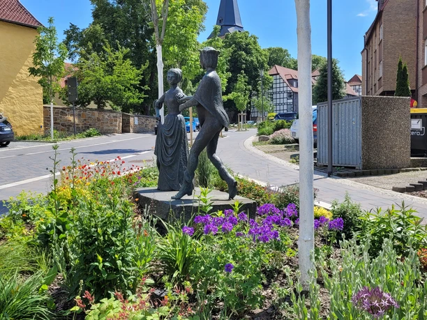 Danzelstätte Lübbecke Bronzeskulptur eines tanzenden Paars inmitten eines bunt blühenden Blumenbeets vor Fachwerkhäusern.