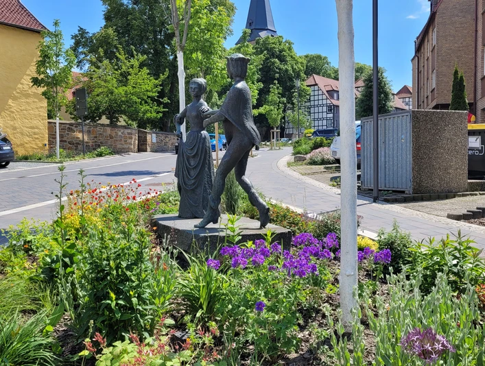 Danzelstätte Lübbecke Bronzeskulptur eines tanzenden Paars inmitten eines bunt blühenden Blumenbeets vor Fachwerkhäusern.