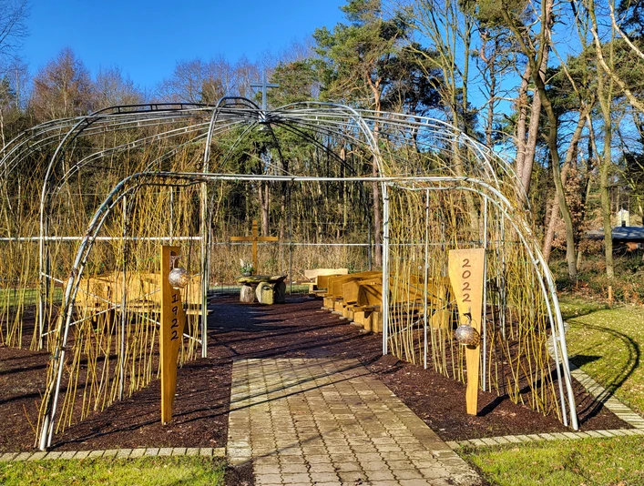 Weidenkirche in Lahn (3) © Naturpark Hümmling e. V..jpg Offene Weidenkirche mit Holzbänken und Kreuz inmitten einer grünen Waldlichtung unter blauem Himmel.