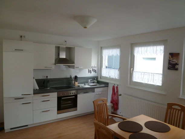 Fewo Höcker_Küche.JPG Alt-Text: Moderne Küche mit weißer Einbauküche, Esstisch, Holzstühlen und hellen Fenstern.