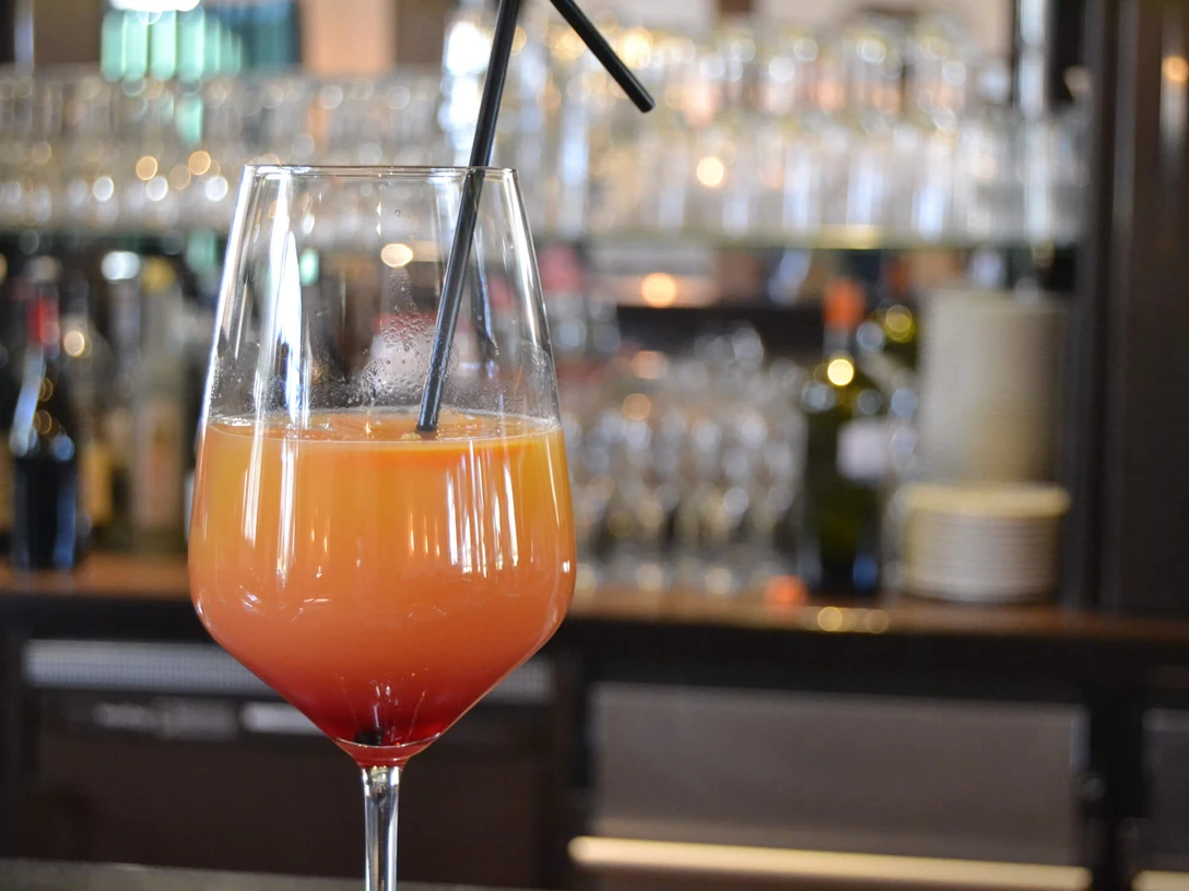 Ein Cocktailglas mit orangem Drink, rotem Boden und Strohhalm vor unscharfem Barhintergrund.