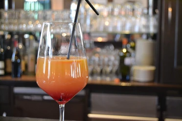 Bar Symbolbild Ein Cocktailglas mit orangem Drink, rotem Boden und Strohhalm vor unscharfem Barhintergrund.