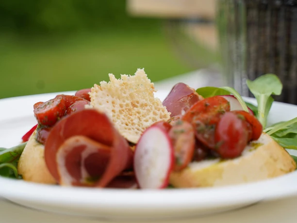 Biergarten Symbolbild Ein Teller mit Bruschetta, belegt mit Schinken, Tomaten, Radieschen und Salat auf einem Holztisch.