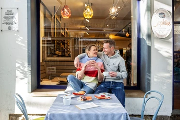 Een lachend stel zit buiten een café te genieten van koffie en cake op een zonnige dag.