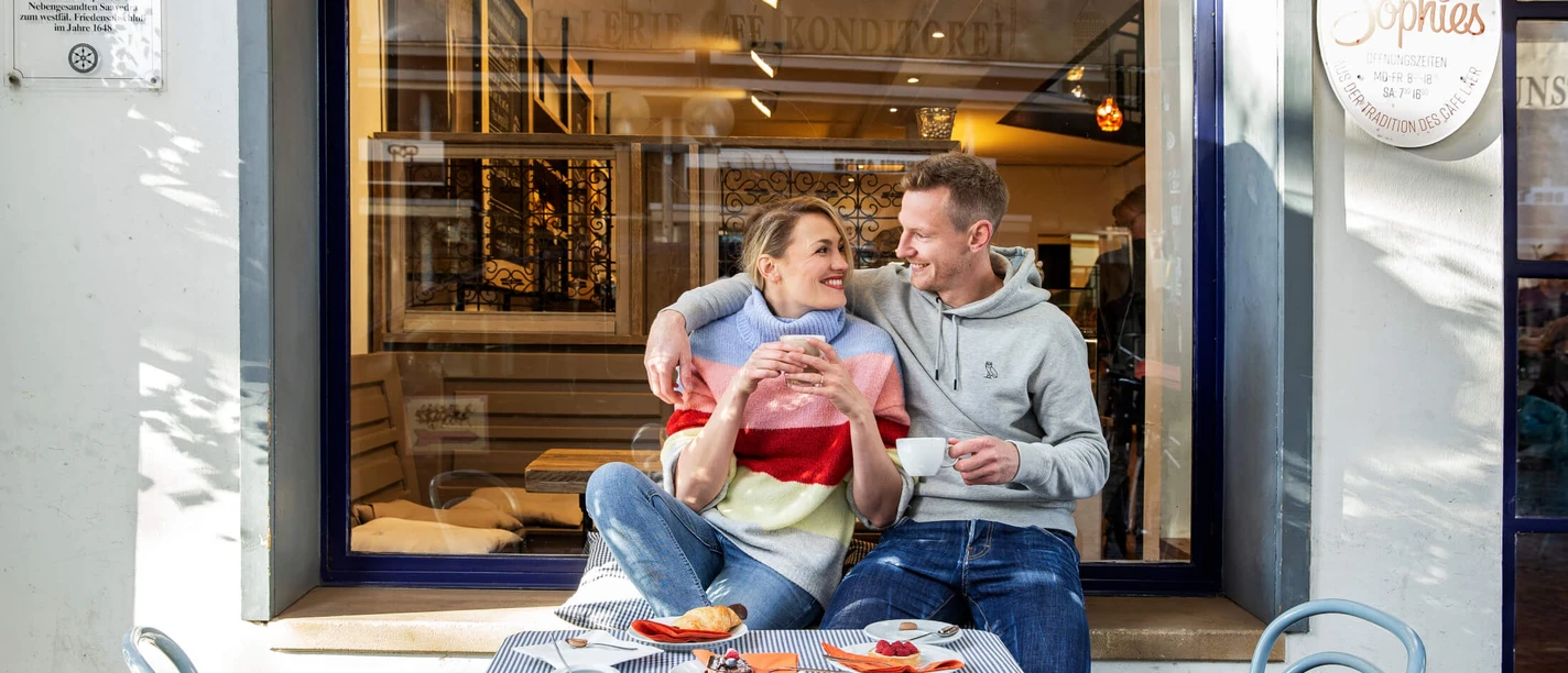 A smiling couple sits outside a café enjoying coffee and cake on a sunny day.