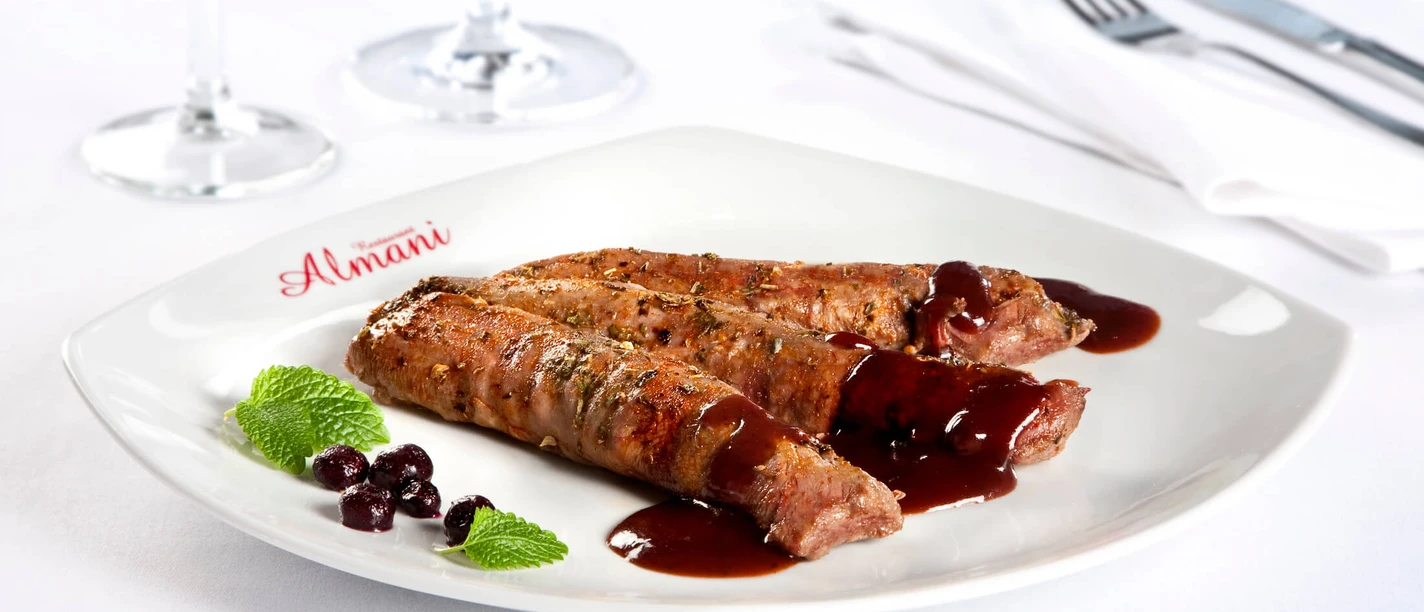 Genuss im Almani Zwei saftige Fleischröllchen mit roter Sauce und Kräutern auf einem Teller mit Almani-Logo.