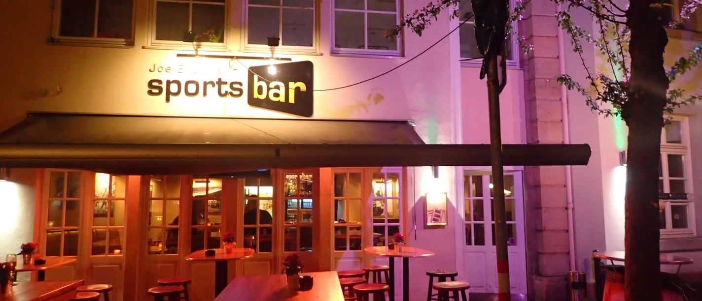 Joe Enochs Sportsbar Sportsbar in Abendstimmung, beleuchtet mit Neonlicht, umgeben von Bäumen mit blühendem Blätterwerk.