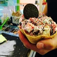 Eisdiele Schlecks in Osnabrück Ein Waffelbecher voller Eis mit bunten Streuseln und Oreo dekoriert, in einer Hand gehalten.