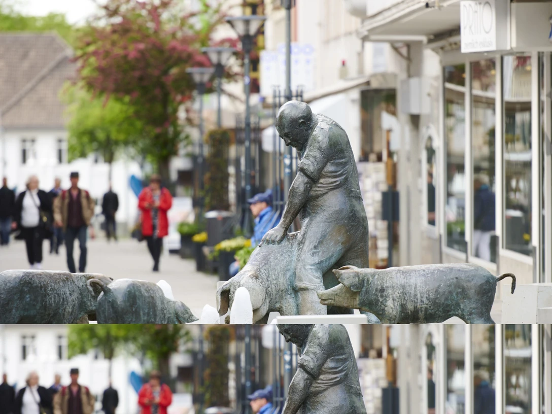 1.png Das Bild zeigt den Schweinebrunnen in Bad Oeynhausen in der Innenstadt. Im Hintergrund sind Menschen zu sehen, die durch die Innenstadt schlendern.