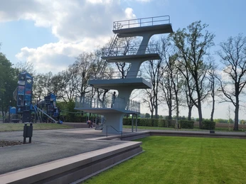 Freibadpark Lübbecke Sprungturm im Freibadpark Lübbecke vor blühenden Bäumen, Himmel mit Wolken und grüne Wiese.