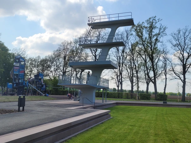 Freibadpark Lübbecke Sprungturm im Freibadpark Lübbecke vor blühenden Bäumen, Himmel mit Wolken und grüne Wiese.