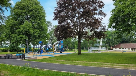 Spielplatz mit blauen Klettergeräten und Rutsche, daneben ein Baum und im Hintergrund ein Sprungturm.