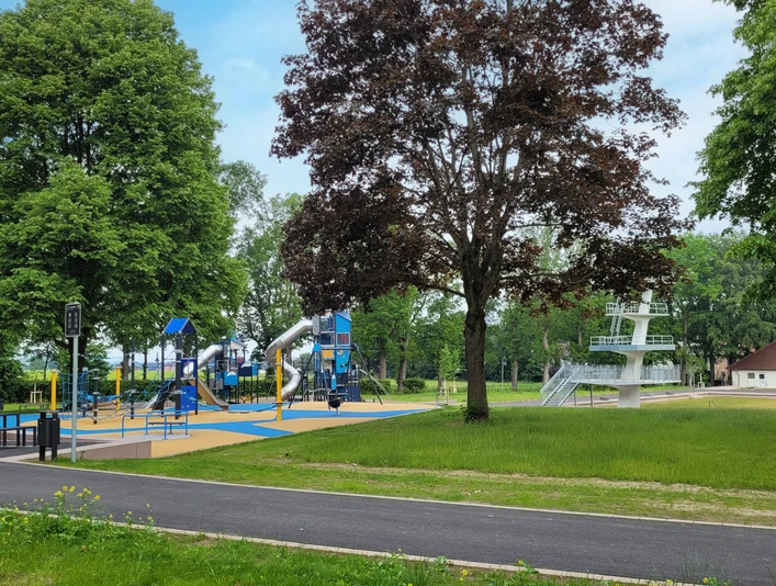 Spielplatz mit blauen Klettergeräten und Rutsche, daneben ein Baum und im Hintergrund ein Sprungturm.