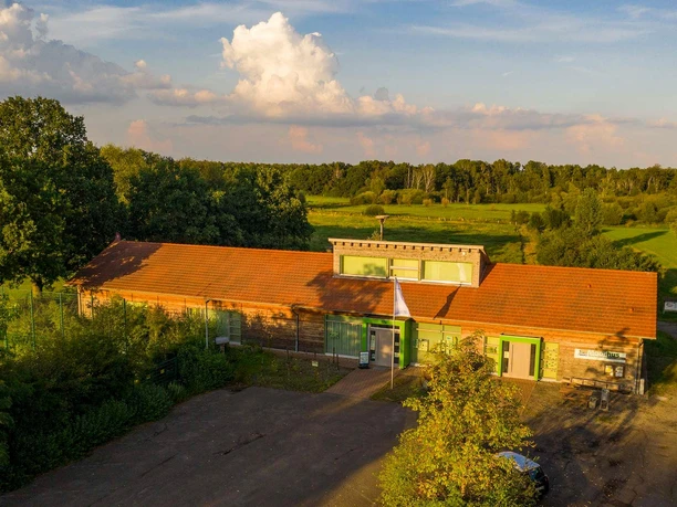 Backsteingebäude mit rotem Dach im Grünen, Sommerwetter; umgeben von Wiesen und Bäumen.
