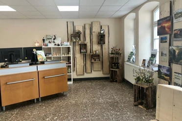 Touristinformation Sebnitz Heller Raum mit Holzmöbeln, Regalen mit Souvenirs und Broschüren, zwei Computermonitoren auf einem Tresen und dekorativen Blumenarrangements.