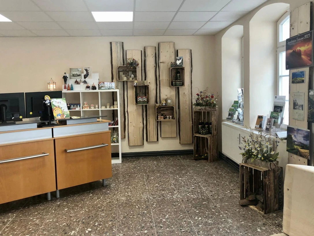 Touristinformation Sebnitz Heller Raum mit Holzmöbeln, Regalen mit Souvenirs und Broschüren, zwei Computermonitoren auf einem Tresen und dekorativen Blumenarrangements.