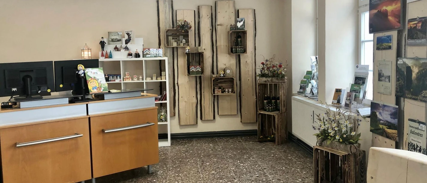 Touristinformation Sebnitz Heller Raum mit Holzmöbeln, Regalen mit Souvenirs und Broschüren, zwei Computermonitoren auf einem Tresen und dekorativen Blumenarrangements.