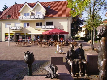 Holter Kirchplatz mit modernem Wohnhaus, Straßencafé, rotem Sonnenschirm und Bronzeskulpturen.