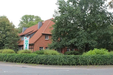 Hotel building with red tiled roof, green hedge and trees in the foreground on a street corner.