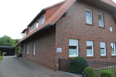 Bakstenen gebouw met zadeldak in het landelijke Uchte, omgeven door bomen en een geplaveide buitenruimte.