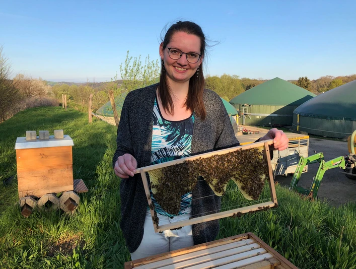 Bien Royal - Imkerei Laube in Langenfeld Eine Person hält ein Bienenwaben-Rahmen in der Nähe von grünen Kuppeln und einer Wiese.