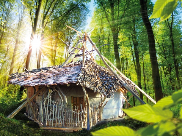 Schamanen-Schaluppe Naturbelassene Hütte im Wald mit Strohdach und Holzverzierungen, von Sonnenstrahlen beleuchtet.