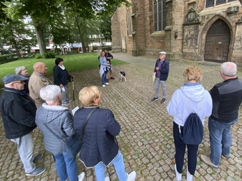 Stadtführungen in Lübbecke_3.jpg Eine Gruppe von Menschen lauscht aufmerksam einem Stadtführer vor einer historischen Kirche in Lübbecke.
