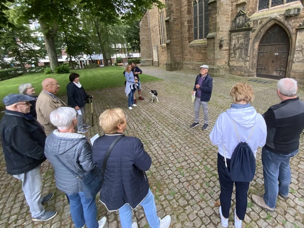 Stadtführungen in Lübbecke_3.jpg Eine Gruppe von Menschen lauscht aufmerksam einem Stadtführer vor einer historischen Kirche in Lübbecke.