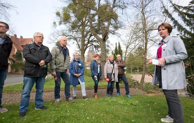 Lübbecke_Stadtführung_Echt oder Ente1_Oliver Krato.jpg Eine Gruppe von Menschen lauscht einer Stadtführerin im Freien, umgeben von Bäumen und historischen Gebäuden.