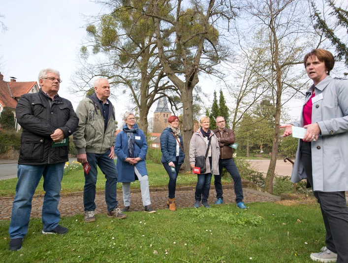 Lübbecke_Stadtführung_Echt oder Ente1_Oliver Krato.jpg Eine Gruppe von Menschen lauscht einer Stadtführerin im Freien, umgeben von Bäumen und historischen Gebäuden.