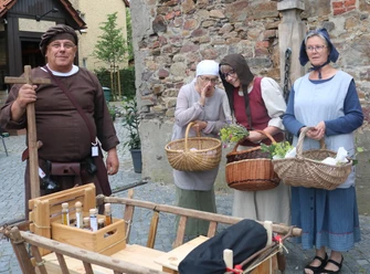 Lübbecke_Stadtführung_Marktgeschehen2_Lübbecke Marketing.JPG Vier Personen in mittelalterlichen Gewändern mit Körben lächeln auf einem Marktplatz in Lübbecke.