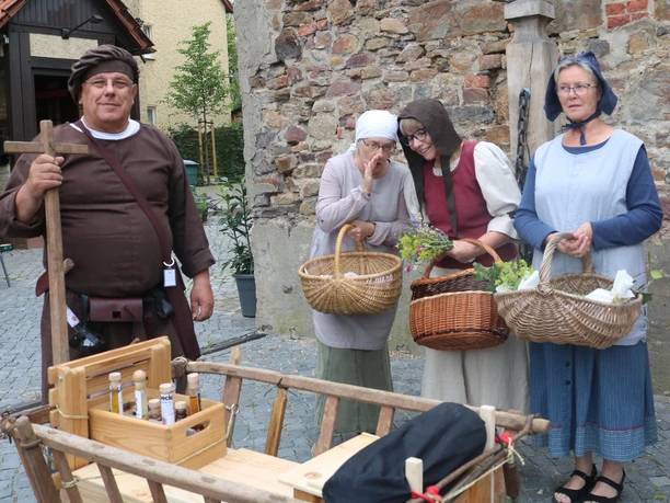 Lübbecke_Stadtführung_Marktgeschehen2_Lübbecke Marketing.JPG Vier Personen in mittelalterlichen Gewändern mit Körben lächeln auf einem Marktplatz in Lübbecke.