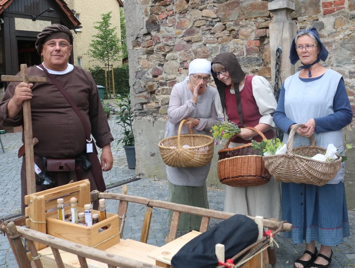 Lübbecke_Stadtführung_Marktgeschehen2_Lübbecke Marketing.JPG Vier Personen in mittelalterlichen Gewändern mit Körben lächeln auf einem Marktplatz in Lübbecke.