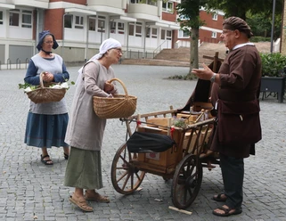 Lübbecke_Stadtführung_Marktgeschehen1_Lübbecke Marketing.JPG Drei Personen in historischer Kleidung beim Gespräch an einem Handwagen auf einem gepflasterten Platz.