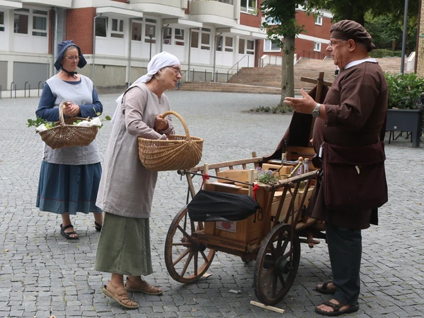 Lübbecke_Stadtführung_Marktgeschehen1_Lübbecke Marketing.JPG Drei Personen in historischer Kleidung beim Gespräch an einem Handwagen auf einem gepflasterten Platz.
