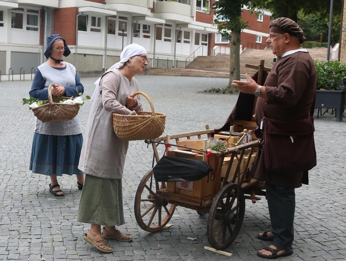 Lübbecke_Stadtführung_Marktgeschehen1_Lübbecke Marketing.JPG Drei Personen in historischer Kleidung beim Gespräch an einem Handwagen auf einem gepflasterten Platz.