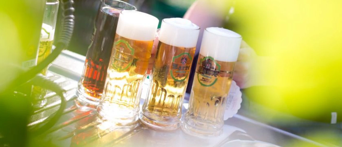 faf_prosit.jpg Vier Bierkrüge mit Schaumkrone stehen auf einem Tresen im Freien, Lichtstrahlen fallen darauf.