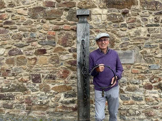 Ein älterer Mann steht neben einem historischen Schandpfahl vor einer alten Steinmauer.