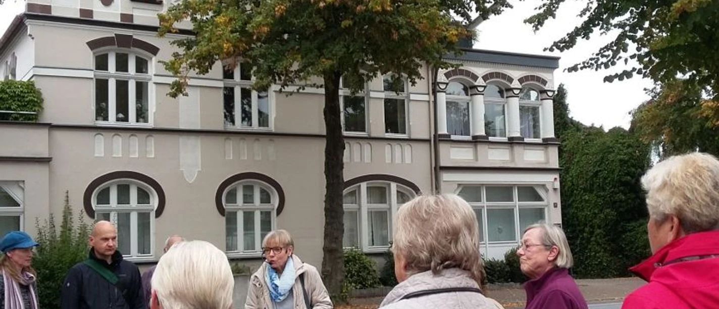 20161003_151528.jpg Eine Gruppe älterer Menschen steht vor einem historischen Gebäude unter einem Baum und hört zu.