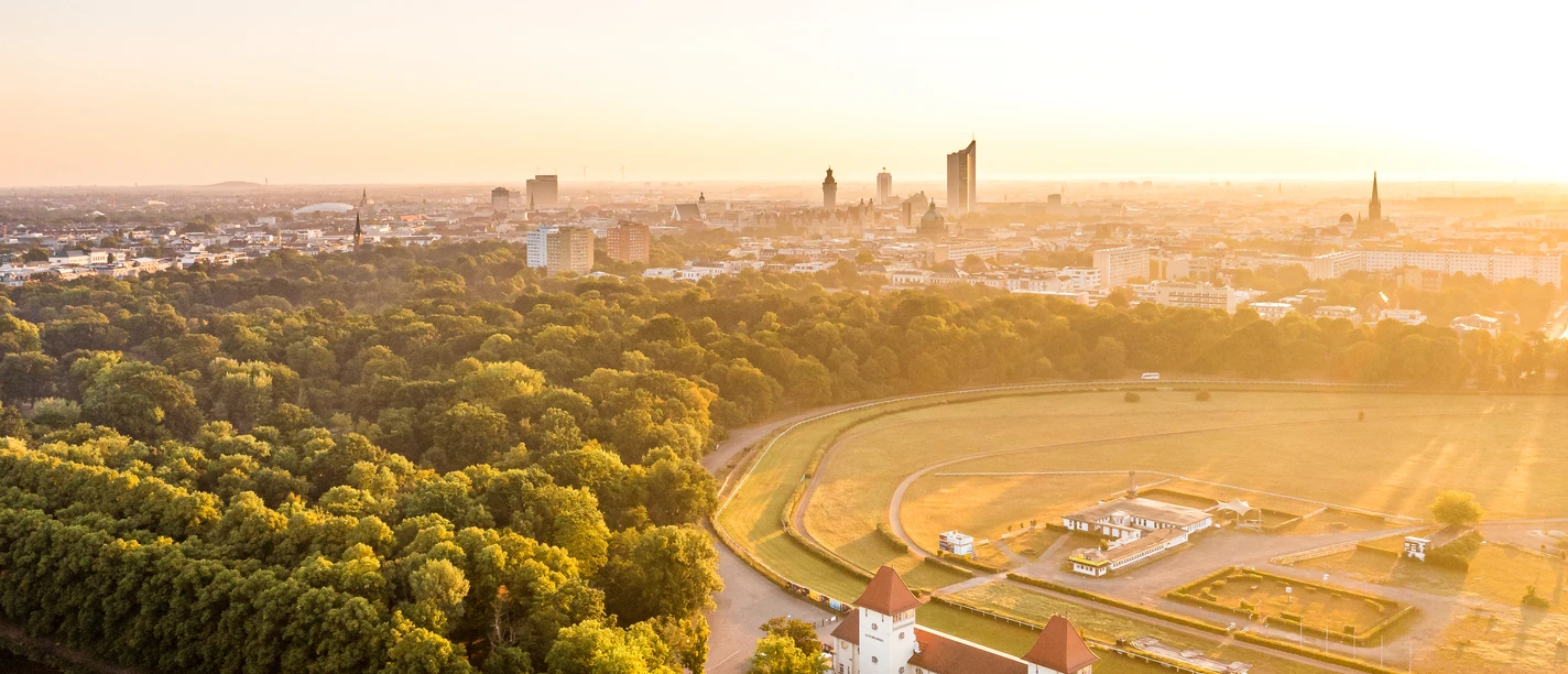 Letecký snímek zachycuje závodní dráhu Scheibenholz při východu slunce a v pozadí panoráma Lipska