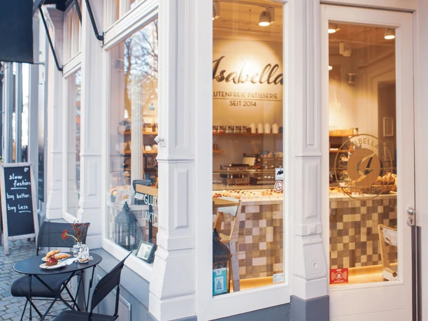 Isabella Glutenfreie Patisserie Außenansicht mit großen Fenstern, die in weißen Rahmeneingefasst sind. Davor steht ein kleiner runder Tisch mit zwei Stühlen. Hinter der gläsernen Eingangstür befindet sich der Innenraum mit einerTheke.