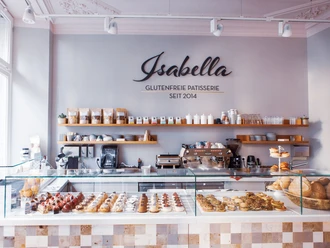 Blick auf eine Theke, hinter deren Glas kleine Gepäckstücke mit Beeren sowie Brotwaren ausgelegt sind. Auf der Wand dahinter steht "Isabella Glutenfreie Patisserie seit 2014" in Großbuchstaben. Darunter ist ein langes Holzbrett an der Wand befestigt, auf dem Gläser, Tassen, Gewürze sowie eine Topfpflanze stehen. Auf zwei kleineren Holzbrettern darunter stehen weitere Tassen und gestapelte Untertassen. Auf dem Küchentisch darunter steht eine große Kaffeemaschine sowie weitere Küchenutensilien.
