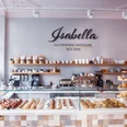 Isabella Glutenfreie Patisserie Blick auf eine Theke, hinter deren Glas kleine Gepäckstücke mit Beeren sowie Brotwaren ausgelegt sind. Auf der Wand dahinter steht "Isabella Glutenfreie Patisserie seit 2014" in Großbuchstaben. Darunter ist ein langes Holzbrett an der Wand befestigt, auf dem Gläser, Tassen, Gewürze sowie eine Topfpflanze stehen. Auf zwei kleineren Holzbrettern darunter stehen weitere Tassen und gestapelte Untertassen. Auf dem Küchentisch darunter steht eine große Kaffeemaschine sowie weitere Küchenutensilien.