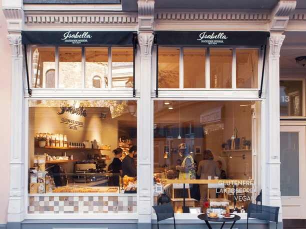 Außenansicht mit einem Tisch und zwei Stühlen im Vordergrund. Durch die gläsernen, großen Schaufenster, von denen auf einem "GLUTENFREI", "LAKTOSEFREI" und "VEGAN" steht, schaut man in den Innenraum des Cafés, in dem eine Frau hinter der Theke zwei Kund*innen bedient. Über jedem der beiden Fenster ist eine kleine Markise ausgefahren, auf der "Isabella Glutenfrei Patisserie seit 2014".