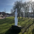 Eine weiße, abstrakte Statue steht auf einem grünen Rasen, umgeben von kahlen Bäumen und Häusern.