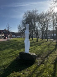 Steinhuder_Nymphe Eine weiße, abstrakte Statue steht auf einem grünen Rasen, umgeben von kahlen Bäumen und Häusern.